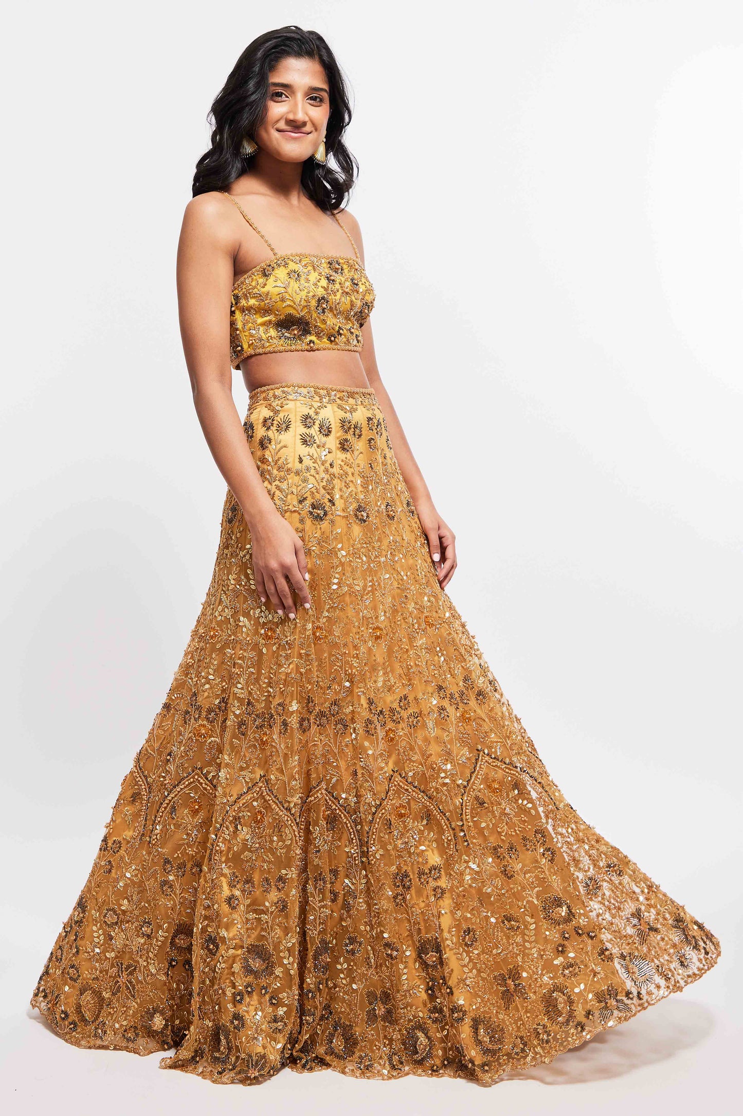 The Hasina Lehenga showcases intricate three-dimensional embroidery, comprising crystal, sequin, and beadwork in an ornate floral pattern adorning both the blouse and skirt. It is elegantly complemented by a matching skinny dupatta with beaded tassels.