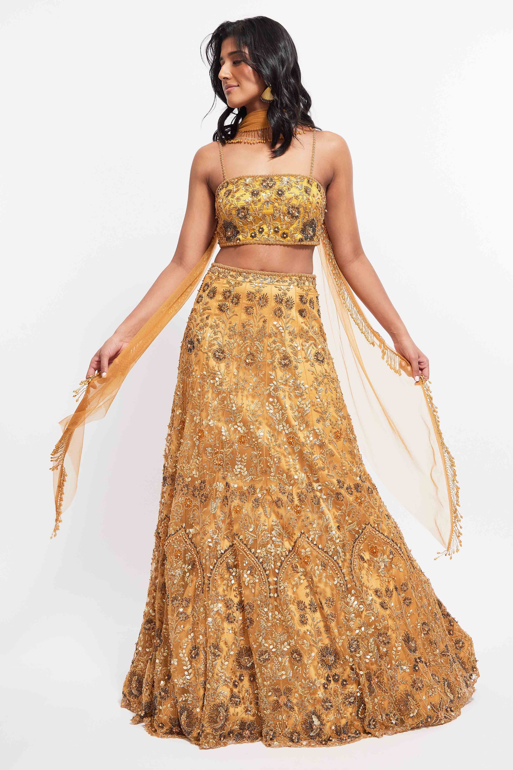 The Hasina Lehenga showcases intricate three-dimensional embroidery, comprising crystal, sequin, and beadwork in an ornate floral pattern adorning both the blouse and skirt. It is elegantly complemented by a matching skinny dupatta with beaded tassels.