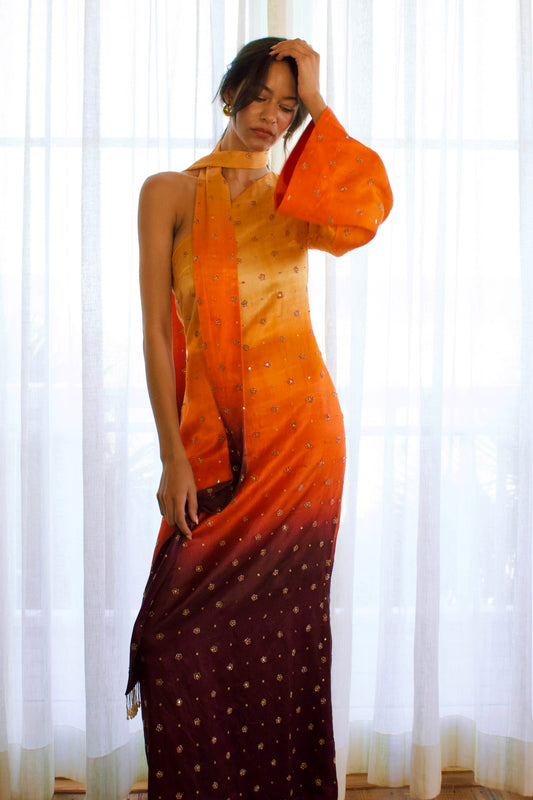 The Amaya Dress features a one-shoulder design with a detachable skinny dupatta. It has a dramatic sleeve and is finished with floral glass beading on ombré fabric in yellow, orange, and brown.