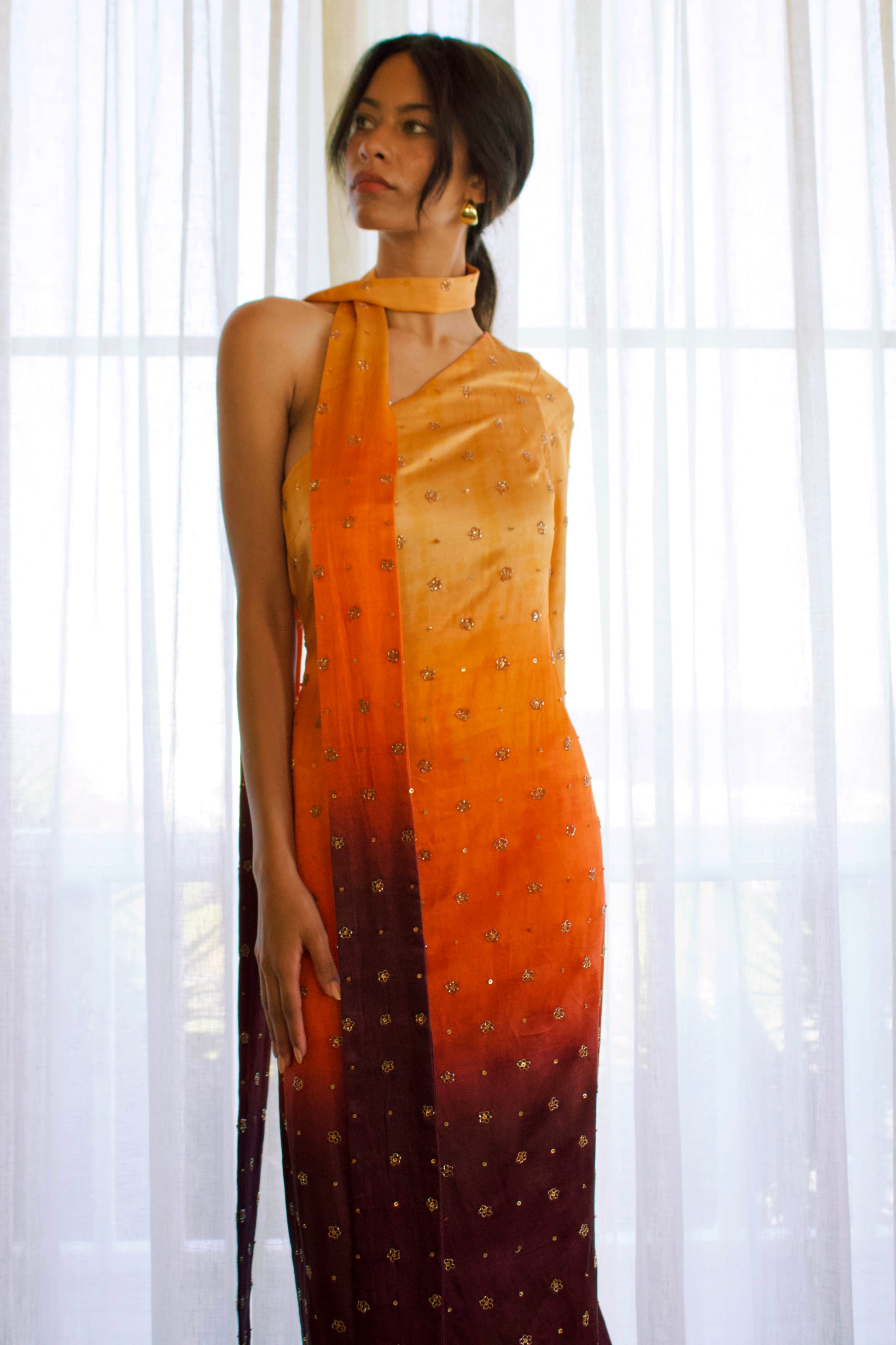 The Amaya Dress features a one-shoulder design with a detachable skinny dupatta. It has a dramatic sleeve and is finished with floral glass beading on ombré fabric in yellow, orange, and brown.