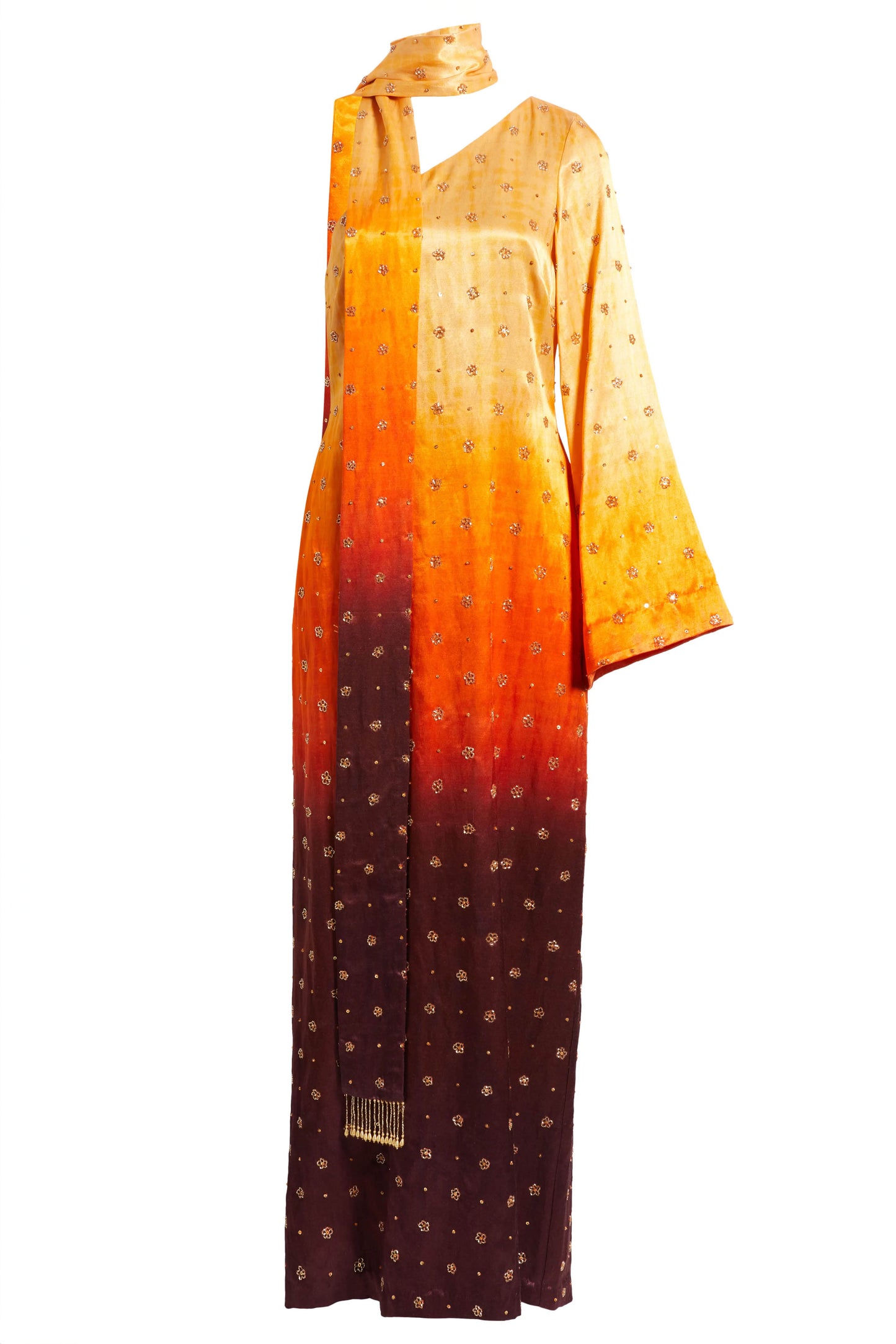 The Amaya Dress features a one-shoulder design with a detachable skinny dupatta. It has a dramatic sleeve and is finished with floral glass beading on ombré fabric in yellow, orange, and brown.