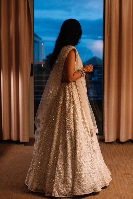 The Rucha Lehenga is a white hand-embroidered lehenga adorned with Swarovski crystals. It features a v-neck blouse with two tie details in the back. It comes with a completely detachable can-can. 