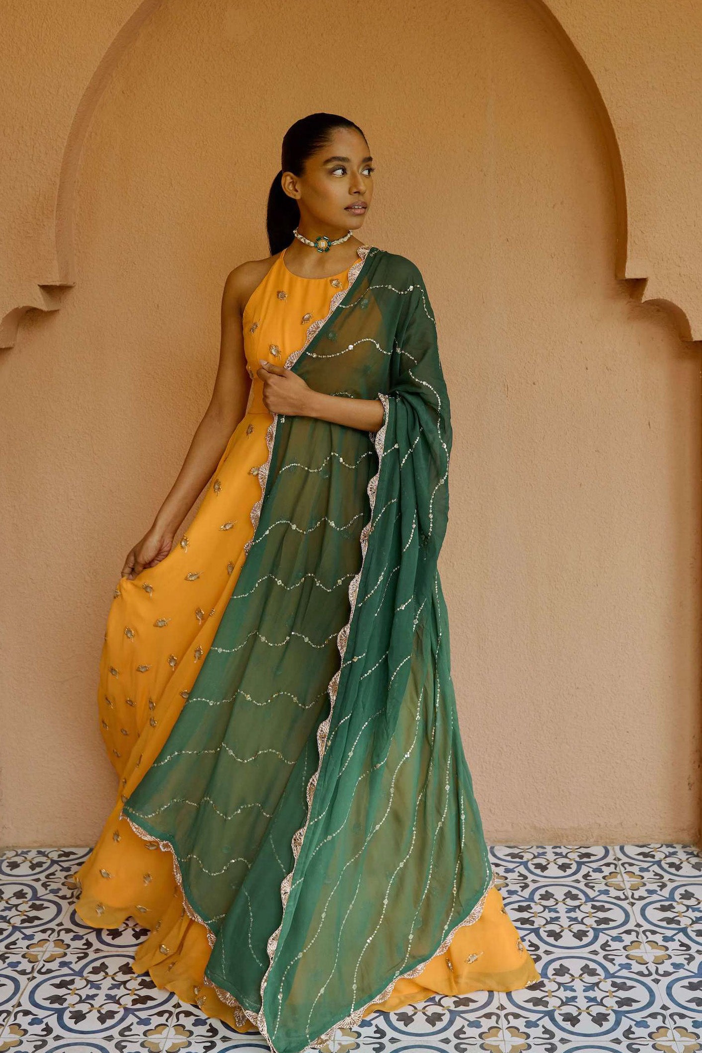 The Nila Anarkali has a halter neck-cut bodice, flowing waistline, and is paired with a contrasting silk crepe dupatta.