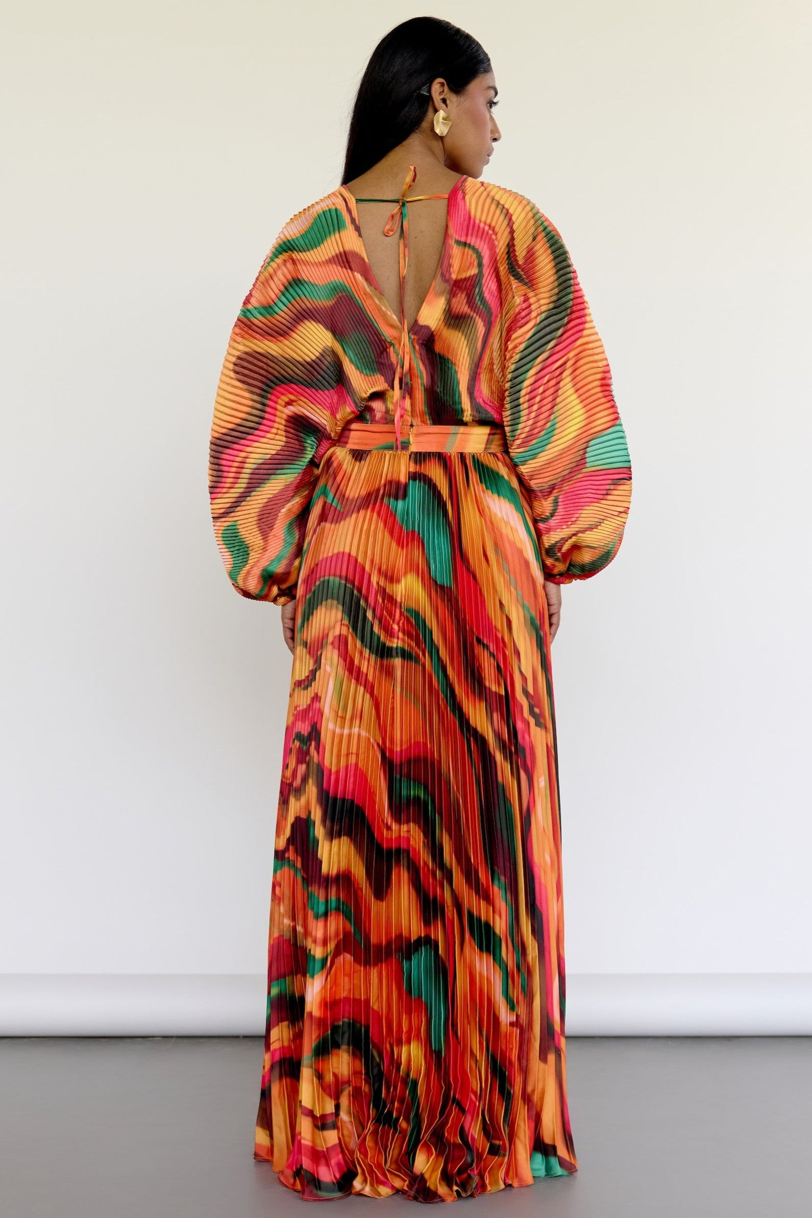 The Sia Dress features a bold abstract print on pleated fabric. Designed with a flattering V-neck, fitted waist, and flowing silhouette, it includes long sleeves, a tie closure at the back, and pockets. Whether you're attending a wedding or heading on a colorful getaway, this dress is an easy statement piece.