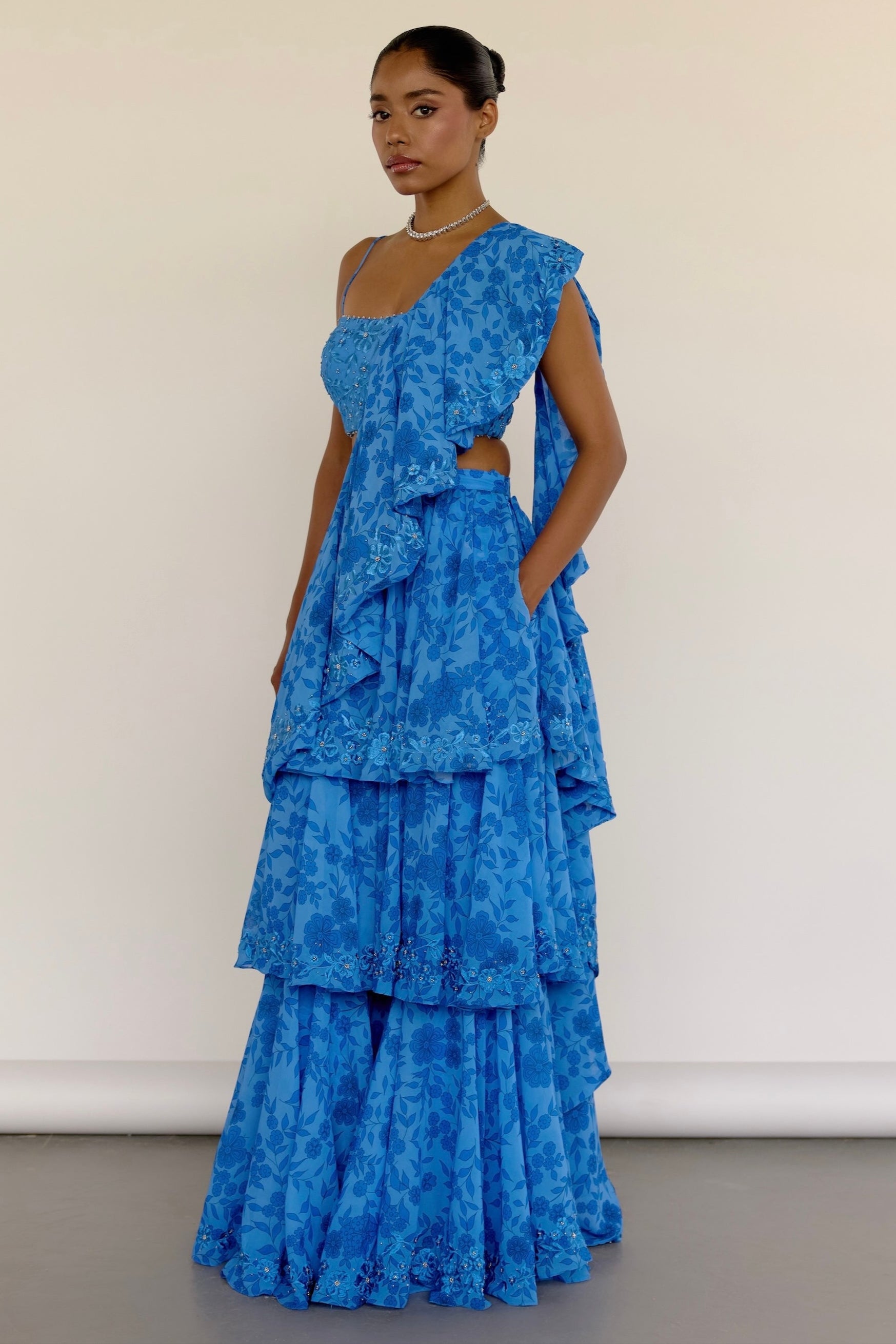 Crafted in a striking blue floral print, the Zaina Palazzo features tiered pants and an embroidered blouse with delicate threadwork and subtle silver highlights. It comes with a detachable shoulder drape that hooks into the pants.